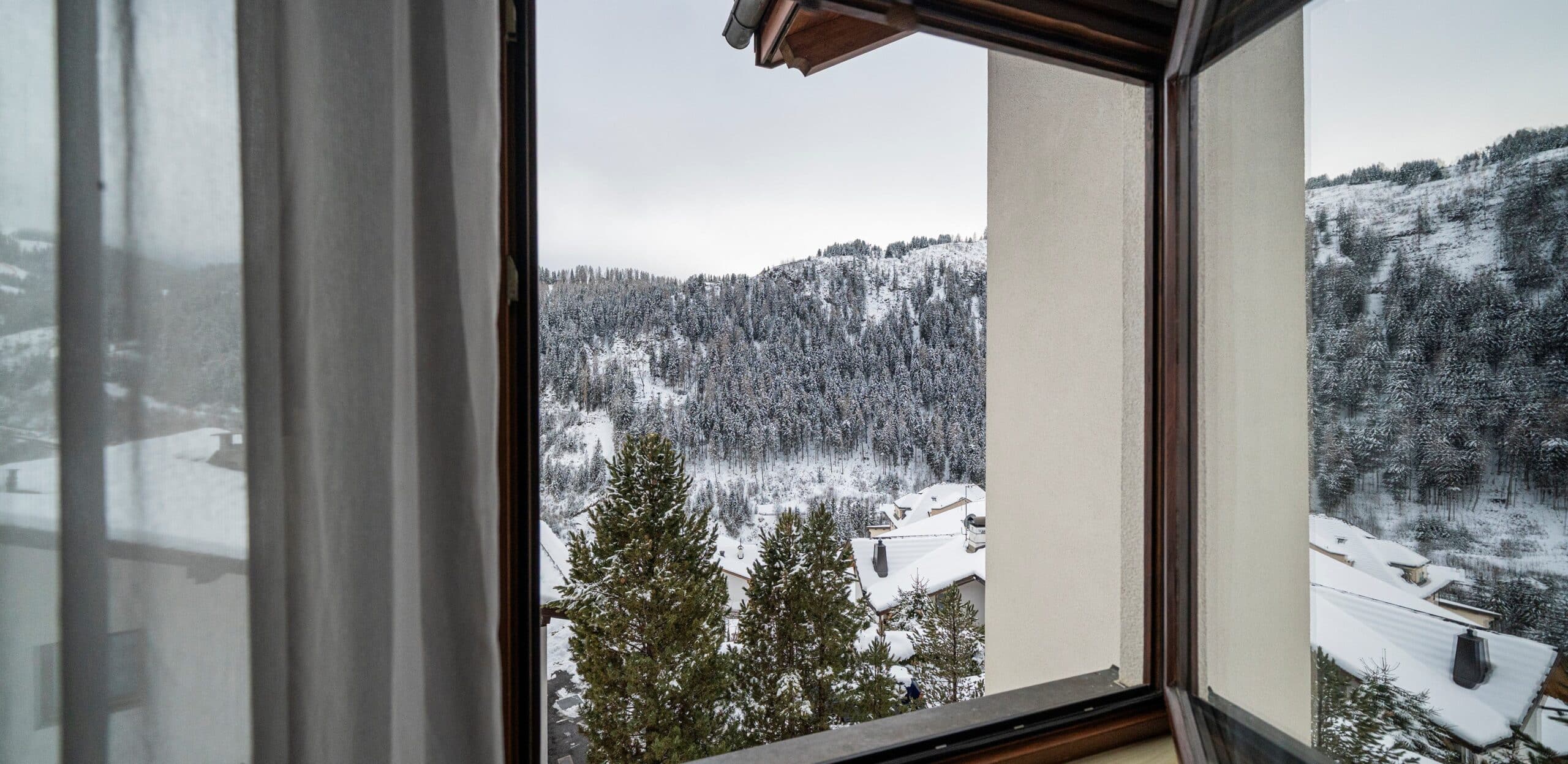 Vista da finestra aperta su tetti innevati, pini in primo piano e monte boscoso coperto di neve