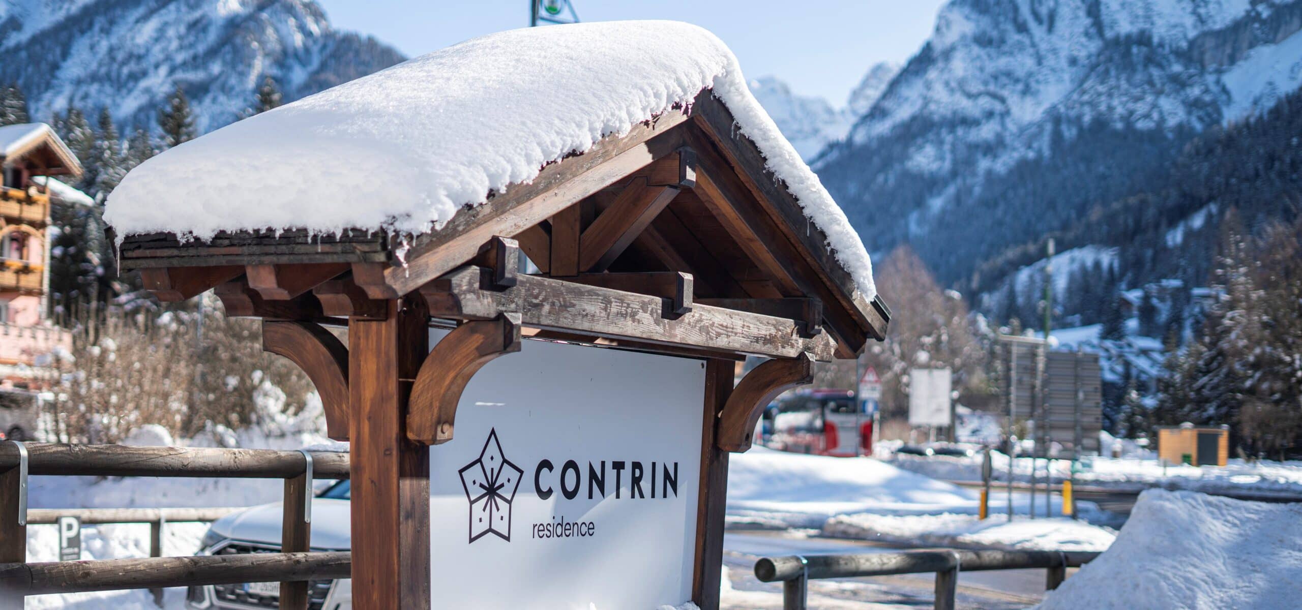 Cartello CONTRIN residence su struttura in legno con tetto ricoperto di neve, montagne sullo sfondo