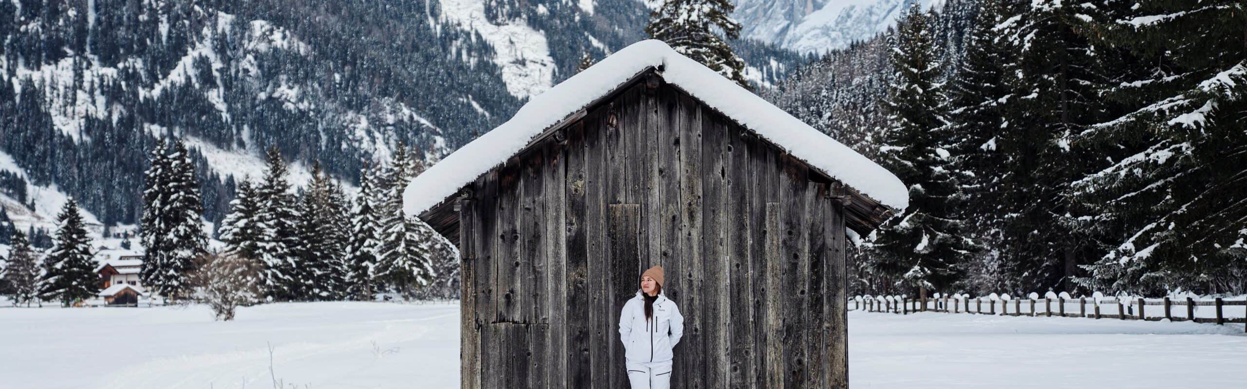 Persona in giacca bianca davanti a un capanno di legno con tetto innevato, pini e montagne sullo sfondo