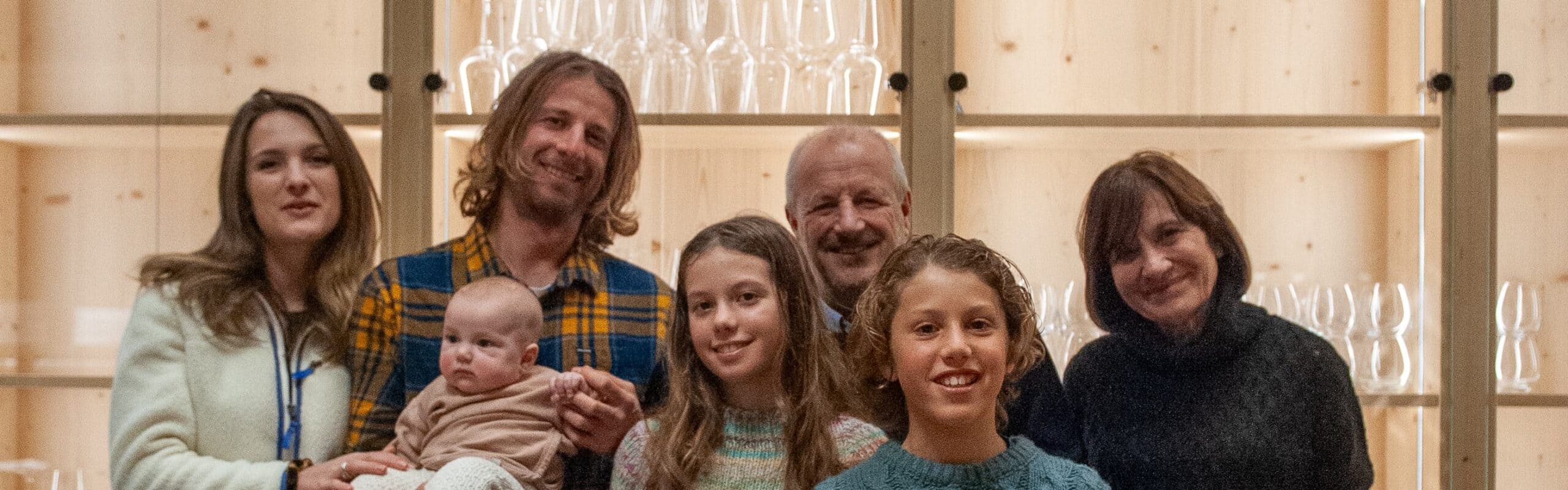 Sette persone, adulti e bambini, uno tiene un neonato, in posa davanti a scaffale di legno con calici