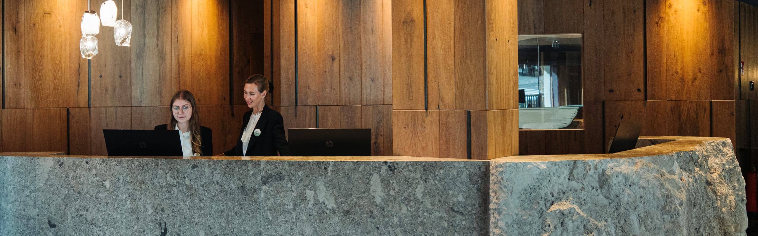 Rezeption mit massiver Steintheke, zwei Mitarbeitende an Laptops vor Holzvertäfelung.